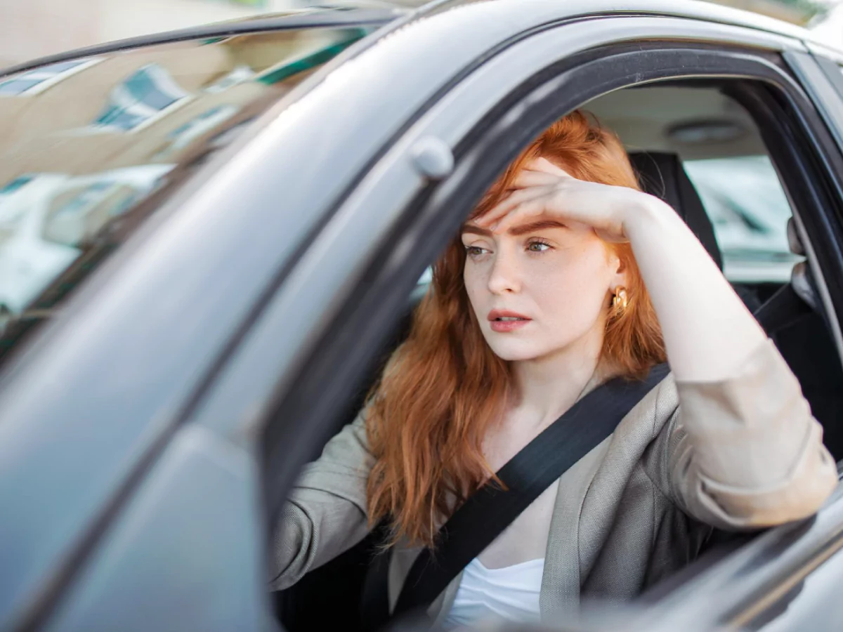 Uma mulher ruiva, usando uma camisa branca e um blazer cinza, está sentada no banco do motorista de um carro. Ela está com uma mão no volante e um braço apoiado na janela aberta, com a mão na testa. Ela olha preocupada para a frente.