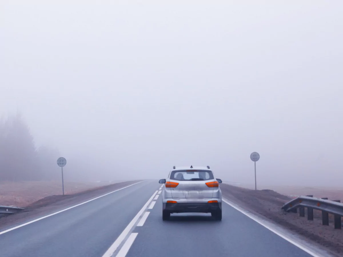 A imagem mostra um carro cinza em uma estrada vazia. Nas margens da estrada é possível ver a forma de duas placas de trânsito, mas a sinalização não é visível. Ao fundo, há a forma de árvores, encobertas por neblina.
