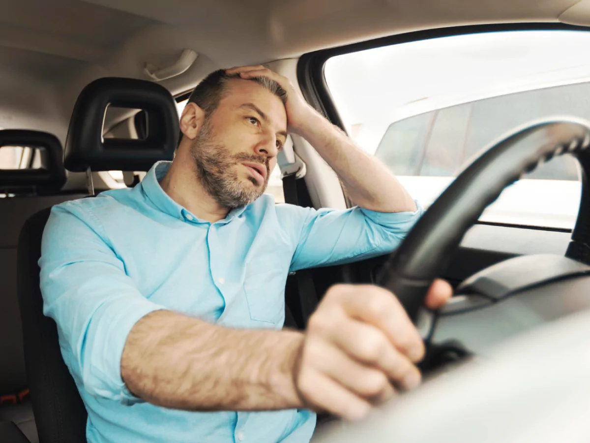 A imagem mostra um homem sentado no banco do motorista de um carro. Ele tem cabelo curto e barba, veste uma camisa azul-marinho e está com o cotovelo apoiado na janela do carro e uma mão nos cabelos. A outra mão segura o volante. O homem tem uma expressão de preocupação.