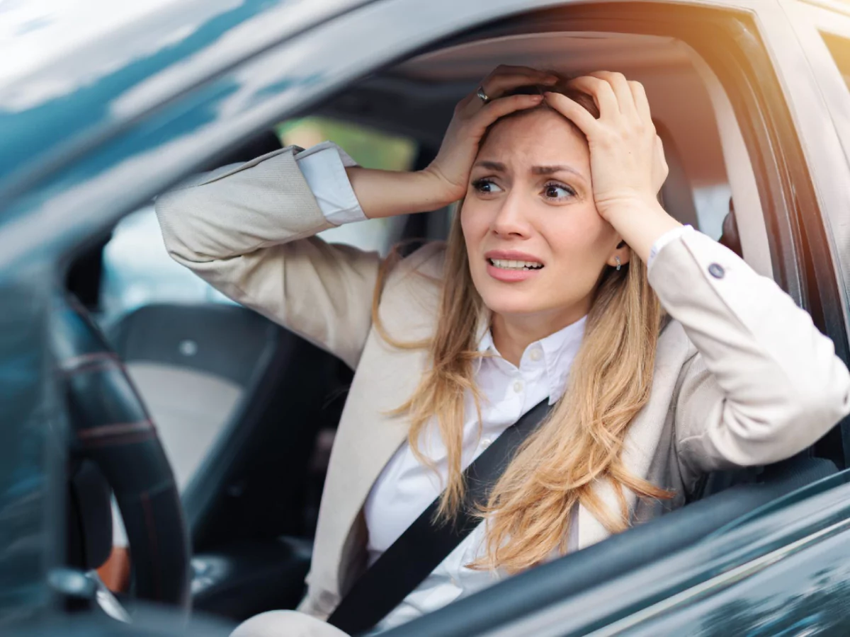 Uma mulher de cabelo loiro, vestindo uma camisa branca e um blazer marrom-claro, está sentada no banco do motorista de um carro. Ela usa o cinto de segurança e está com as duas mãos na cabeça e uma expressão de desespero.