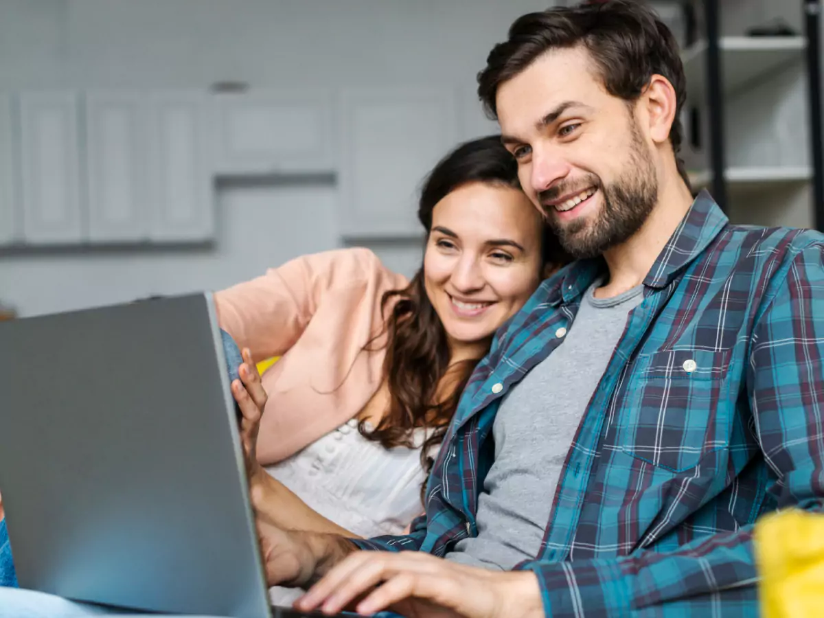 Um homem de camiseta cinza e camisa azul e uma mulher com casaco rosa observam, sorridentes, a tela de um notebook cinza.