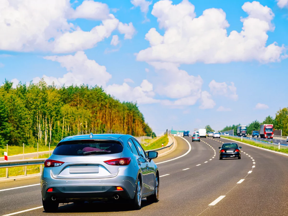 A imagem destaca um carro azul em uma rodovia. Vários veículos estão na estrada. A via é ladeada por vegetação alta e árvores. A parte superior da imagem é ocupada por um céu azul e diversas nuvens brancas.