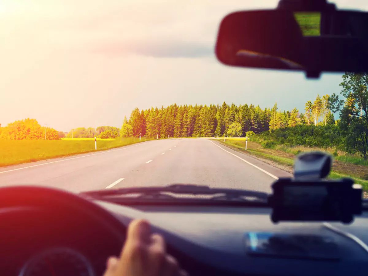 A imagem mostra o painel de um carro, uma mão ao volante e um espelho retrovisor. Pela janela, uma estrada vazia, com árvores ao fundo e nuvens no céu. A iluminação laranjada sugere um pôr-do-sol.