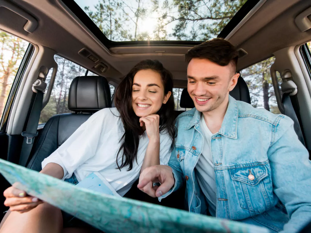 Mulher e homem, ambos dentro de um automóvel, visualizam o que seria um mapa. Detalhe para o teto solar do carro e a vegetação do lado de fora.