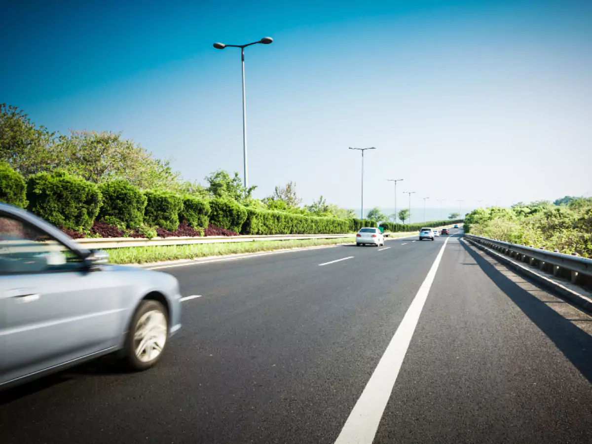 Rodovia de mão única com três carros em movimento. Vegetação nas laterais, postes com luminárias e céu azul compõem a paisagem.