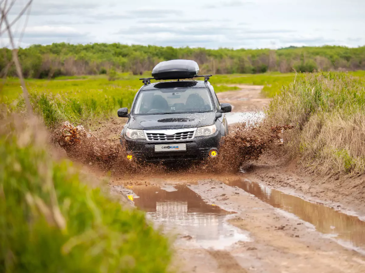 Carro trafega por via não asfaltada, repleta de barro e poças d'água. Nas laterais, há uma vegetação rasteira. Ao fundo, há uma floresta densa.