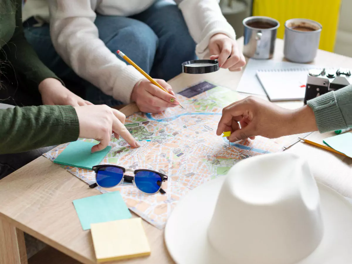 Mãos sobre mapa apontando localidades. Sobre a mesa há um chapéu branco, óculos azuis, post-its e canecas de metal. Uma das pessoas segura um lápis e uma lupa.