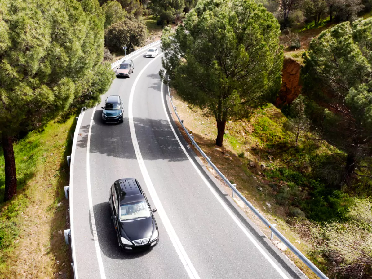 A imagem mostra uma rodovia vista de cima. Quatro carros transitam na mesma direção. A estrada é cercada por árvores e vegetação.