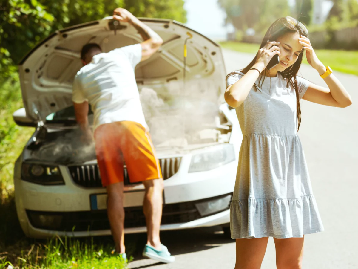 A imagem mostra, em primeiro plano, uma mulher de vestido cinza fazendo uma ligação no celular. Ao fundo, um homem de camisa branca e calção laranja olha para o motor de um carro branco, envolto em fumaça, estacionado na beira de uma estrada.