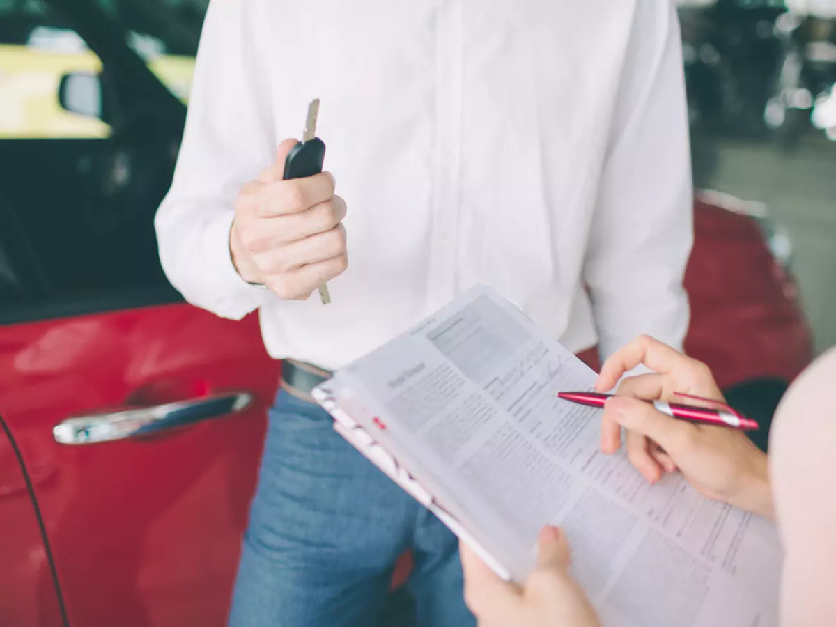 A imagem mostra um homem de camisa branca e calça azul segurando as chaves de um carro. Duas mãos seguranm folhas de um contrato e uma caneta vermelha. Ao fundo, um carro vermelho.