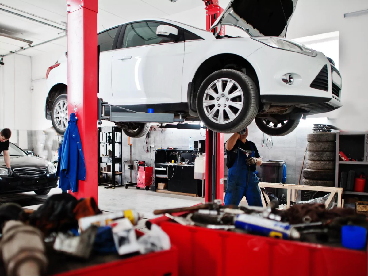 A imagem mostra uma oficina mecânica. Um carro branco está suspenso no ar, enquanto um mecânico, vestindo um macacão azul, trabalha na parte inferior do carro. Ao fundo, um carro preto, no chão, tem seus faróis dianteiros examinados.