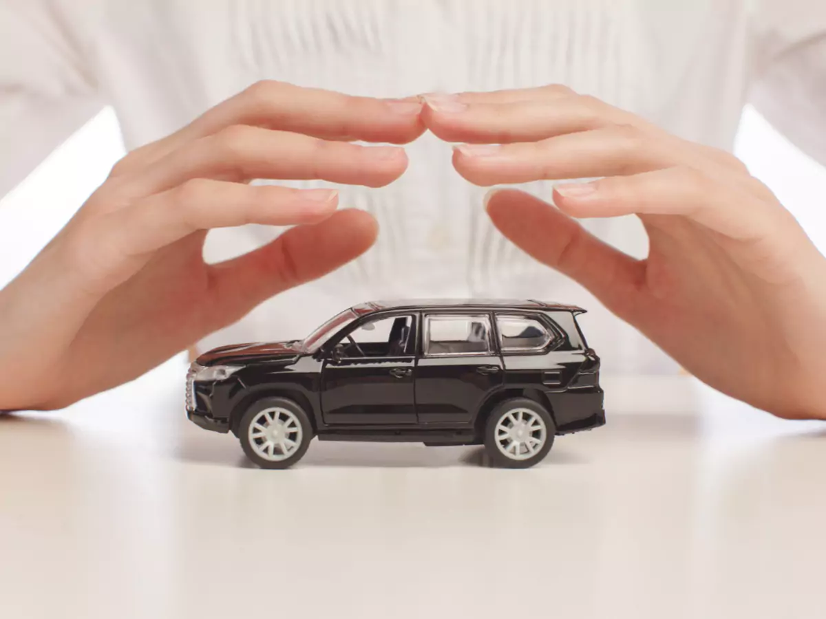 Uma miniatura de um carro preto aparece em destaque, com um par de mãos formando um triângulo sobre o veículo. Ao fundo, o torso da pessoa que faz este gesto de proteção está coberto por uma camisa branca.