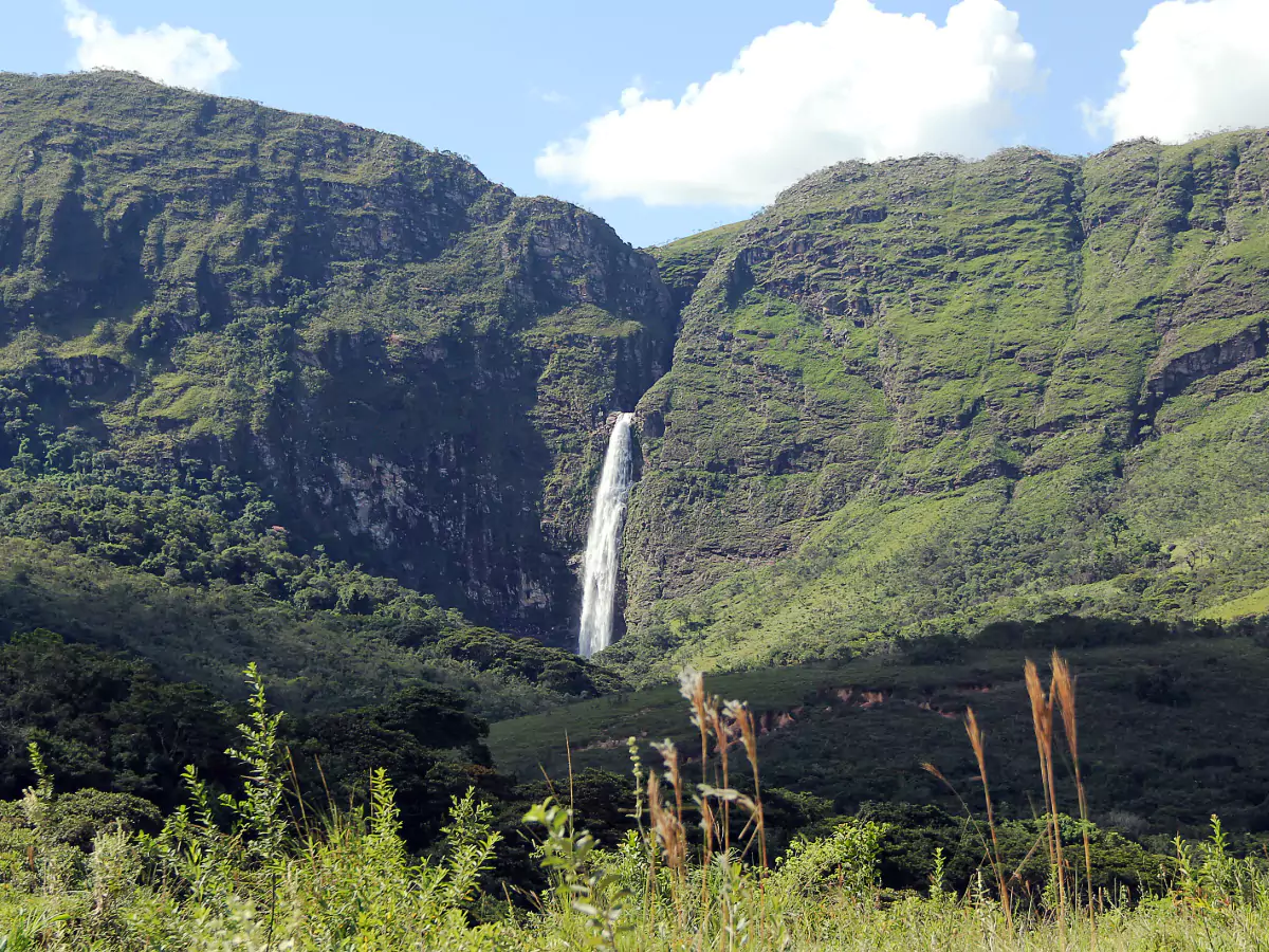 Gigantesca montanha com cachoeira formando um paredão na Serra da Canastra. A planície é composta por pequenos elevados, e toda a imensidão é coberta por ampla vegetação, contrastando com o céu azul e poucas nuvens.
