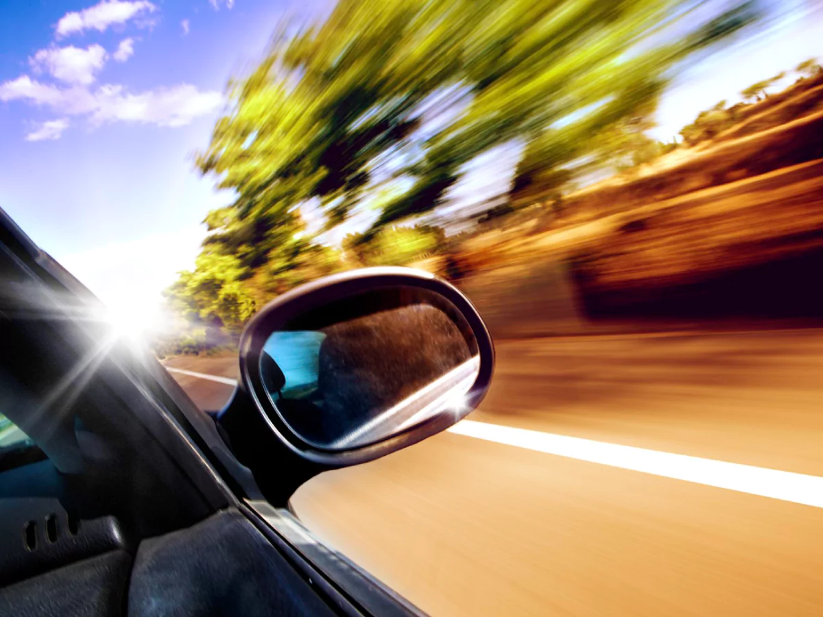 A imagem mostra a visão da janela de um carro preto em alta velocidade. A janela está aberta e o foco está no espelho retrovisor. Ao fundo, a estrada está borrada, assim como as árvores que ladeiam a via. O céu azul tem algumas nuvens brancas.
