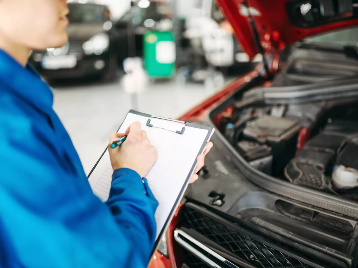 Um homem vestido de azul segura uma prancheta e uma caneta verde. Ele olha para o motor de um carro laranjado, que está com o capô aberto na sua frente. Ao fundo, um carro preto desfocado.