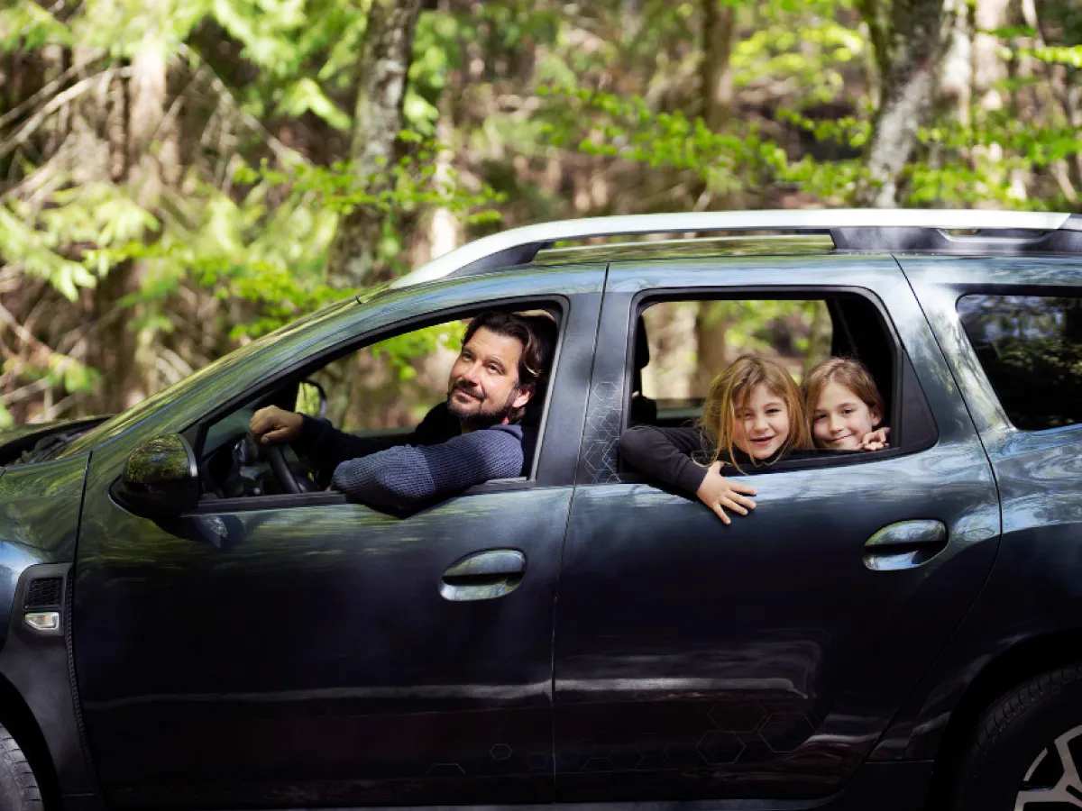 A imagem mostra a lateral de um carro preto. No lugar do motorista, um homem com um braço apoiado na janela aberta e uma mão no volante olha para a câmera. No banco traseiro, duas crianças também encaram a câmera, sorridentes. Ao fundo, é possível ver árvores e vegetação verde.