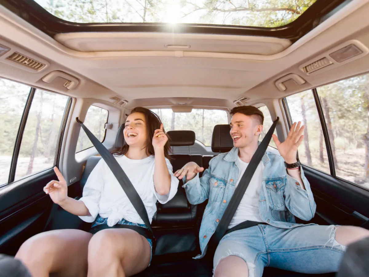 A imagem mostra o interior de um carro com janelas amplas e teto solar. É possível ver árvores do lado de fora do veículo. No interior, dois jovens estão sentados, usando o cinto de segurança. Na esquerda, uma mulher de cabelo comprido usando um short azul e uma camisa branca. Na direita, um homem usa calções e casaco jeans e uma camisa branca.