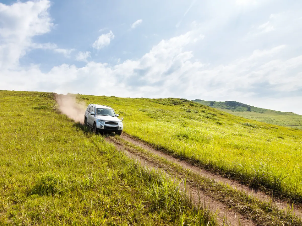 A imagem mostra um veículo branco avançando por uma estrada de terra. O carro levanta uma nuvem de poeira atrás de si. A estrada é ladeada por vegetação verde baixa. Ao fundo, é possível ver o céu azul repleto de nuvens brancas.