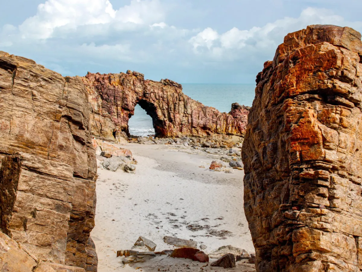 A imagem mostra um trecho da rota da Costa do Sol Poente. É possível ver várias formações rochosas de diferentes cores de marrom em uma praia. Ao fundo, o mar azul e o céu coberto de nuvens brancas.