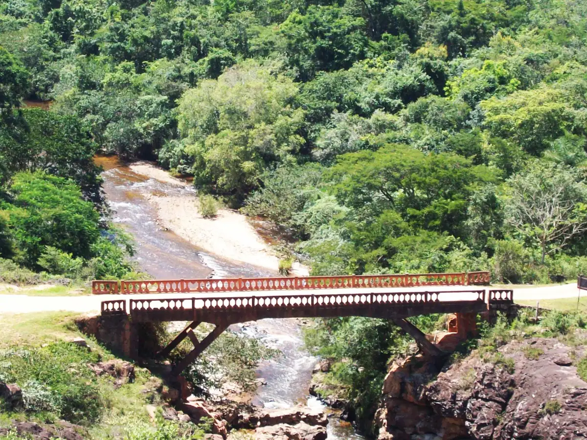 A imagem mostra uma visão aérea de um trecho da Estrada Real. Uma ponte de madeira sobre um rio. É possível ver bancos de areia nas margens, a encosta rochosa em que a ponte se apoia e uma floresta densa ao fundo.