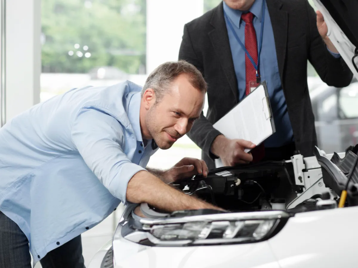 Na imagem, dois homens olham para o interior do capô de um carro branco. Um deles está inclinado para dentro do capô, com as mãos dentro do compartimento do motor. Ele usa uma camisa azul e uma calça jeans. O segundo homem está em pé, usando um terno preto, camisa azul e uma gravata vermelha, segurando uma prancheta.