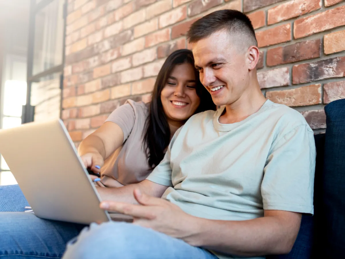 Um homem e uma mulher estão sentados. Ele tem um laptop no colo e sorri para a tela. A mulher aponta algo na tela do computador e também sorri.