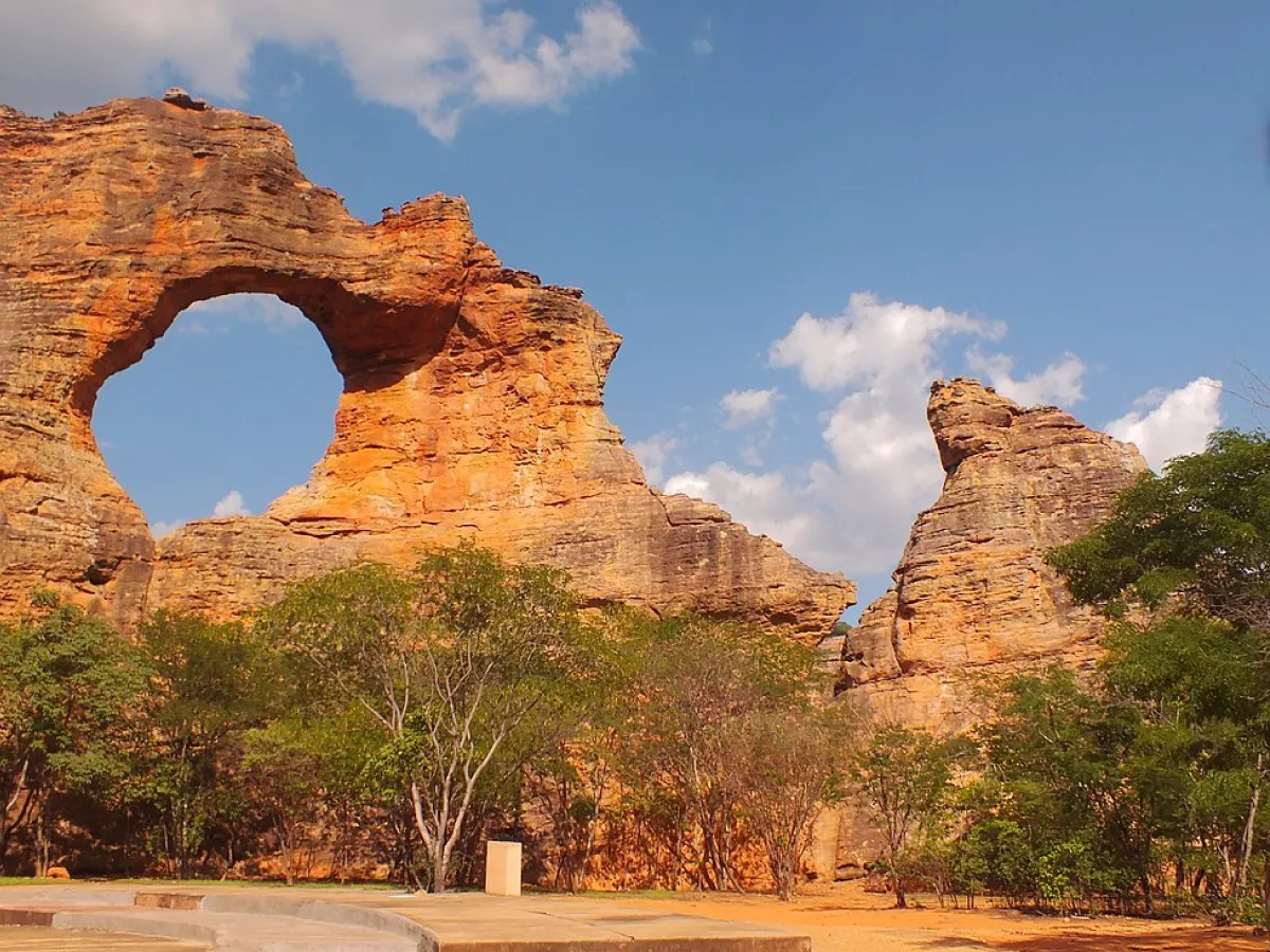 A imagem mostra uma paisagem do Parque da Serra da Capivara. É possível ver formações rochosas altas e árvores com a folhagem verde. Ao fundo, o céu azul e algumas nuvens brancas.