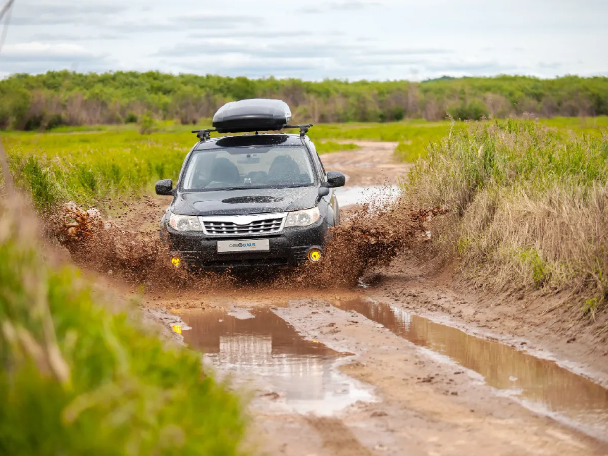 Um carro preto, com um bagageiro superior, avança por uma estrada de terra alagada. Água com barro se levanta nas laterais do veículo. A estrada é ladeada de vegetação verde. Ao fundo, é possível ver uma vegetação alta e fechada, e o céu azul acinzentado com nuvens.