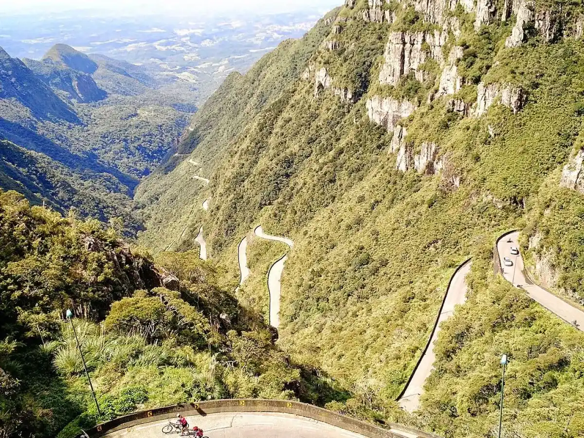 A imagem mostra uma vista aérea da Serra do Rio do Rastro. É possível ver montanhas cobertas de vegetação verde e uma estrada que se estende por toda a paisagem. Do lado direito, é possível ver alguns veículos transitando pela via. Na parte inferior da imagem, alguns ciclistas seguem a rota.