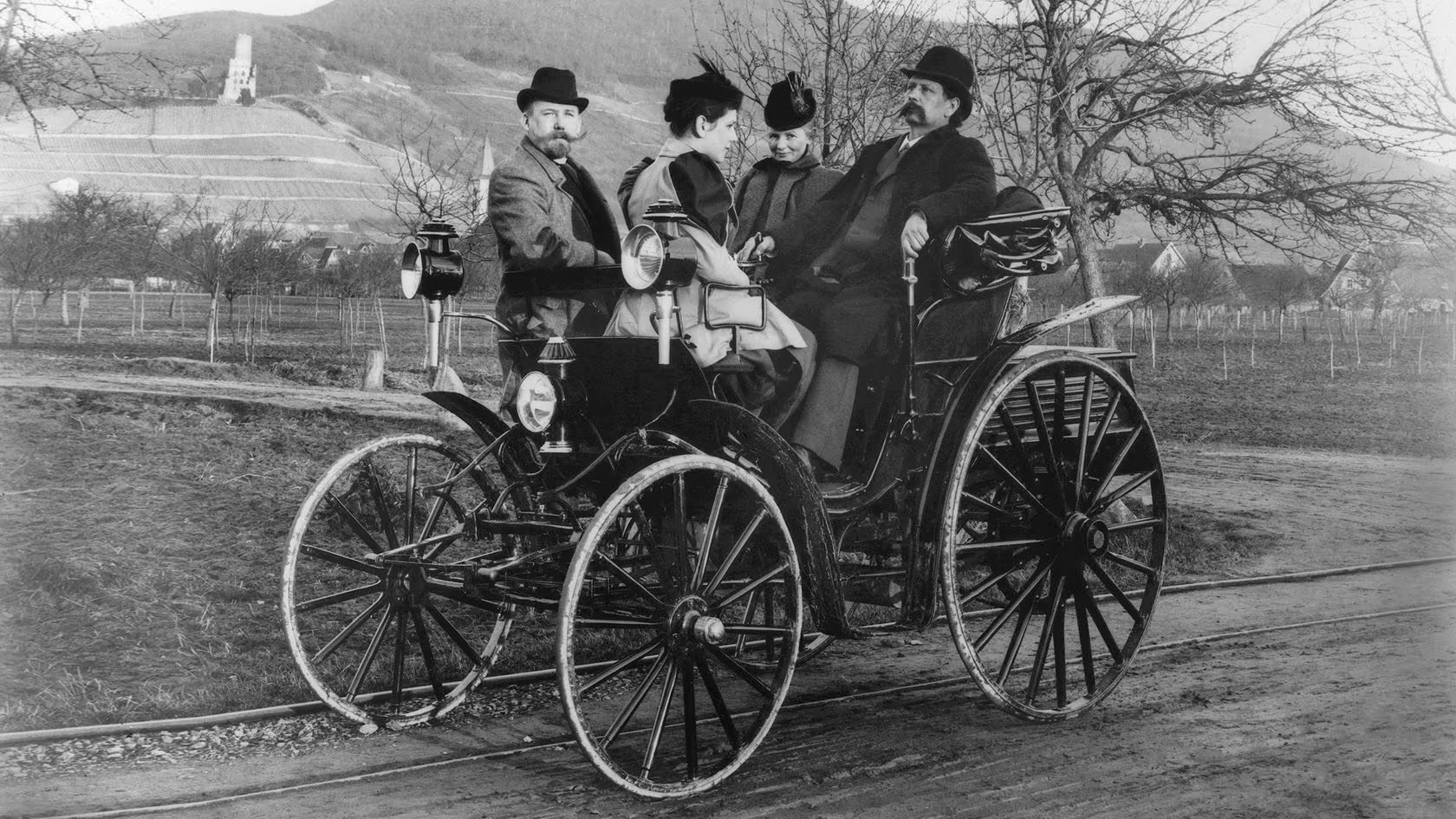 A imagem histórica, em preto e branco, mostra Karl e Bertha Benz acompanhados de sua filha Clara e Fritz Held em um dos carros inventados por Karl. O veículo é totalmente aberto, lembrando uma carruagem, com quatro rodas altas e finas. O carro está parado em uma estrada de terra.