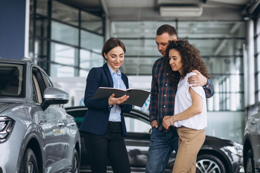 Um homem e uma mulher observam um caderno segurado pela atendente da locadora de carros em que eles estão.