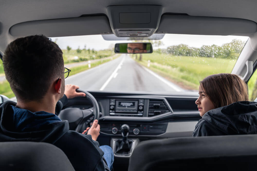Um homem e sua filha estão no banco da frente do carro. Ele dirige com atenção em uma estrada reta com gramas em volta.