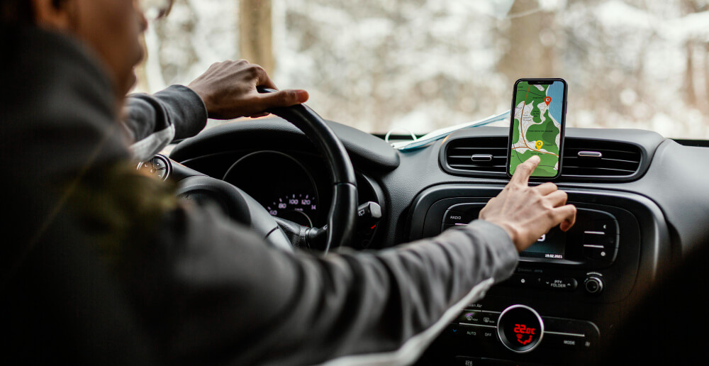 Homem com jaqueta cinza dirige enquanto ajusta o GPS no celular preso ao suporte no painel do carro.